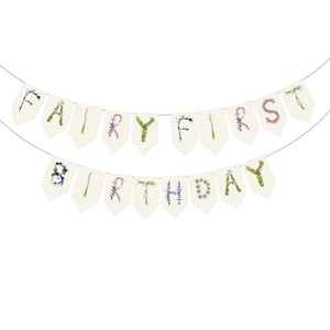 May include: A banner with the words "FAIRY FIRST BIRTHDAY" spelled out on individual cream-colored pennants. The letters are formed with colorful flowers and greenery, strung on a thin wire against a white background.