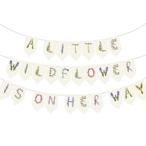 May include: A banner with three rows of pennants spelling out "A LITTLE WILDFLOWER IS ON HER WAY." Each letter is decorated with floral designs in various colors, including purple, green, and pink. The pennants are cream-colored and strung on a thin white string.