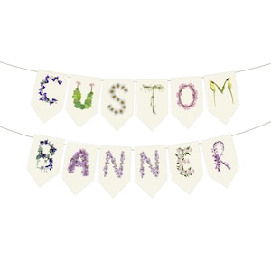 May include: A banner spelling "CUSTOM BANNER" with floral letters. Each letter is formed by arranging different types of flowers in various colors, including purple, green, and white, on individual cream-colored pennant flags suspended by a thin string.