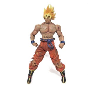 May include: A muscular action figure of a character with spiky yellow hair, a bare chest with battle-worn markings, orange pants with a blue sash, blue wristbands, and black boots with yellow trim, standing in a tense, combat-ready pose.