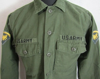Puede incluir: Una camisa de manga larga color verde oliva con cierre de botones, dos bolsillos en el pecho con solapas abotonadas y el texto U.S. ARMY bordado en negro sobre cada bolsillo. Cada hombro muestra un parche con un emblema de águila amarilla.
