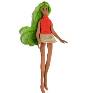 May include: A doll with long, bright green hair, a tan complexion, and painted facial features. The doll wears a sleeveless, textured orange top with a high neckline and a short, plain beige skirt. The limbs are slender and elongated in design.