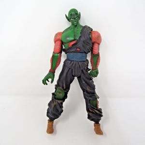 May include: A green-skinned humanoid action figure with pointed ears, wearing a tattered dark purple sleeveless tunic, a blue sash, and dark purple pants with rips at the knees. The figure has pinkish-red textured arm segments and brown boots.
