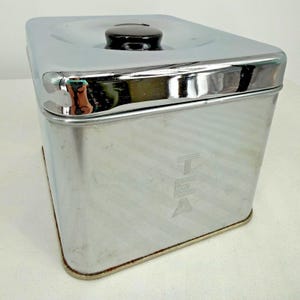 May include: A square, chrome-finished metal tea canister with rounded corners and a reflective surface. The canister features a black knob handle on the lid and the word TEA embossed vertically in a stylized font on the front side of the container.