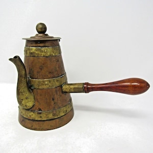 May include: A copper coffee pot featuring three horizontal brass bands, a curved brass spout, a matching brass lid with a spherical finial, and a long, smooth, dark brown wooden handle extending horizontally from the side of the vessel.