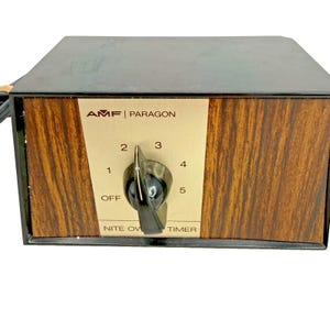 May include: An AMF Paragon Nite Ov timer with a black rectangular casing and a wood grain front panel. The center features a beige control plate with a black dial, marked with settings from OFF to 5, and the text AMF PARAGON NITE OV TIMER.