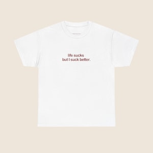 Könnte beinhalten: Weißes T-Shirt mit dem roten Text "life sucks but I suck better." Das Shirt hat einen Rundhalsausschnitt und kurze Ärmel. Der Text ist mittig auf der Vorderseite des Shirts platziert.