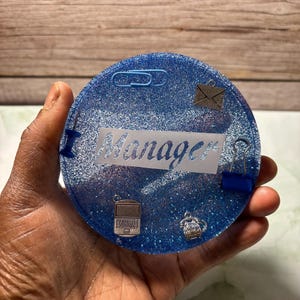 May include: A round, blue glitter coaster with the word "Manager" in white. The coaster is decorated with small charms of office supplies, including a laptop, envelope, and paperclip.
