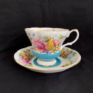 Puede incluir: Una taza de té y un platillo blancos con un diseño floral y de frutas. La taza tiene una banda turquesa en el medio y un borde dorado. El platillo tiene un diseño similar con una banda turquesa alrededor del borde.