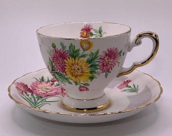 May include: White teacup and saucer set with a floral design featuring pink and yellow flowers and green leaves. The cup and saucer have gold trim. The teacup has a curved handle.