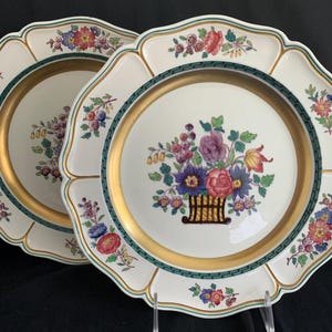 May include: Two white porcelain dinner plates with a gold rim and a green and gold border. The centre of each plate features a colourful floral design with a basket of flowers.