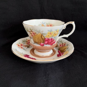 Puede incluir: Una taza de té y un platillo blancos con un patrón floral que presenta peras, cerezas y flores amarillas. La taza y el platillo tienen un borde dorado.