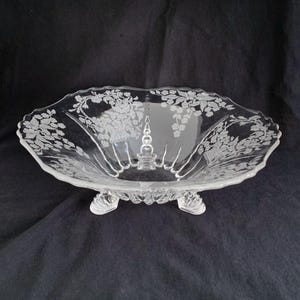 May include: A clear glass pedestal bowl with a floral etched design and three feet. The bowl is ideal for serving fruit, snacks, or desserts.