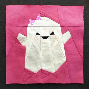 Op de afbeelding: Een roze vierkant van stof met een witte geestenapplicatie. De geest heeft een roze strik met witte stippen en een zwart gezicht met witte ogen en een zwarte mond.