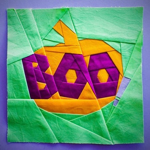 Può includere: Un quadrato di tessuto verde con un'applicazione in tessuto viola e giallo della parola "BOO" in un font stilizzato.