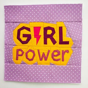 Puede incluir: Una tela morada con lunares blancos presenta una aplicación de tela amarilla con las palabras "GIRL POWER" en letras rosas y moradas. Las letras están hechas de múltiples piezas de tela cosidas juntas.