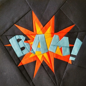 Puede incluir: Un bloque de colcha de tela negra con un diseño de estallido de estrella rojo, naranja y azul. El estallido de estrella tiene la palabra "BAM!" escrita en tela azul.