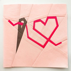 Puede incluir: Un cuadrado de tela rosa con una forma de corazón rosa delineada en tela rosa brillante. También se puede ver una tira de tela marrón en la esquina superior izquierda.