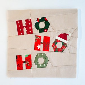 Puede incluir: Un panel de tela beige con un diseño de patchwork con cuadrados de tela roja, verde y blanca. Los cuadrados están dispuestos para deletrear "HO HO HO" con un gorro de Papá Noel en la "O".