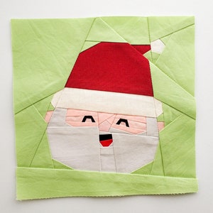 Peut inclure: Un visage de Père Noël en patchwork de tissu vert. Le motif est composé de différentes pièces de tissu de couleurs différentes, dont du rouge, du blanc et du rose. Le visage sourit avec une bouche rouge et des yeux noirs.