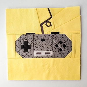 May include: A yellow fabric quilt block with a black and white patterned video game controller design. The controller is in the center of the block and is surrounded by yellow fabric.