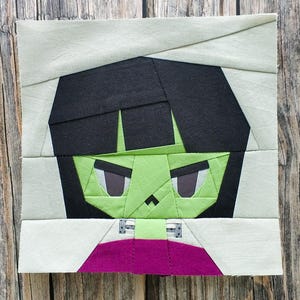 May include: A fabric block featuring a stylized green monster face with black hair and gray eyes. The monster's face is framed by light gray fabric, with a purple collar. The design is made of various fabric pieces sewn together.