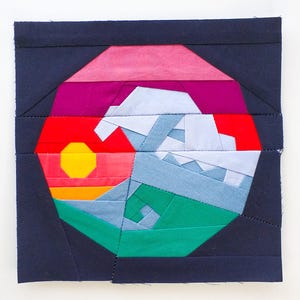Puede incluir: Una pieza de arte textil cuadrada con un diseño de paisaje montañoso. La obra presenta un sol rojo, naranja y amarillo, montañas azules y terreno verde y azul, todo dentro de un borde azul oscuro. La pieza está hecha de tela cosida.