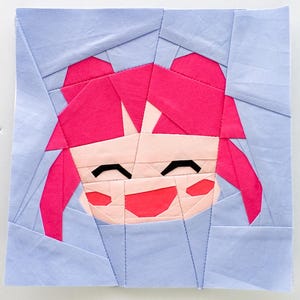 Puede incluir: Un bloque de tela cuadrado que representa un personaje de estilo anime sonriente, con cabello rosa y piel clara. La cara del personaje está hecha de varias piezas de tela, sobre un fondo azul claro. El bloque es probablemente parte de un edredón u otro proyecto textil.