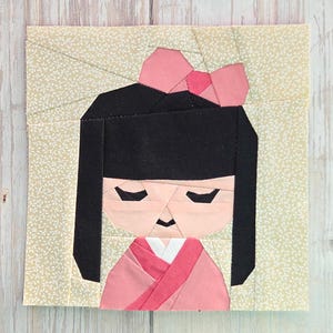 Könnte beinhalten: Ein Stoffkunstwerk mit einem stilisierten Puppen-Design. Die Puppe hat schwarzes Haar, eine rosa Schleife und einen rosa-weißen Kimono. Der Hintergrund ist ein hellbeige Stoff mit einem dezenten Blumenmuster. Das Gesicht der Puppe besteht aus geometrischen Formen.
