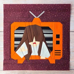 May include: A fabric quilt block featuring an orange television set with a dark brown background speckled with white dots. Inside the screen, a figure with long brown hair covering their face wears a light blue garment, set against a gray striped background.