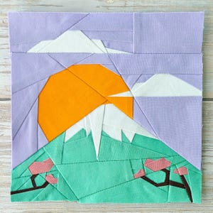 Peut inclure: Une œuvre d'art en tissu représentant un paysage de montagne avec un soleil orange, des nuages blancs et une montagne enneigée. La scène comprend des collines vertes avec des fleurs de cerisier roses sur un ciel lavande.