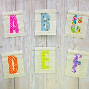 Puede incluir: Seis cuadrados de tela con las letras A a la F aplicadas. Las letras son de varios colores y patrones, incluyendo lunares y estampados florales. El fondo es de color beige claro, sobre una superficie de madera clara.