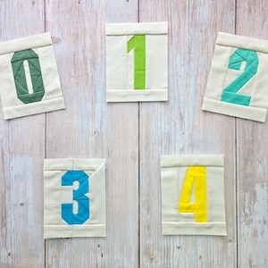 May include: Five fabric blocks displaying the numbers 0 to 4. Each block features a white background with a different coloured number: green, lime green, teal, blue, and yellow. The blocks are arranged on a light wood surface.