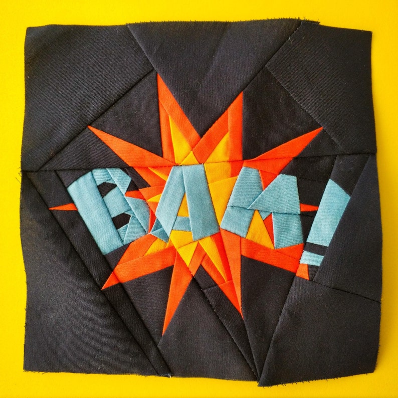May include: A black fabric quilt block with a red, orange, and blue starburst design. The starburst has the word "BAM!" written in blue fabric.