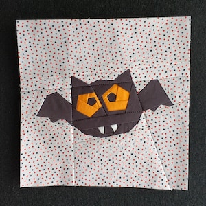 May include: A patchwork fabric square with a bat design. The bat is dark brown with orange eyes and white fangs. The wings are spread. The background fabric is white with a pattern of red and black polka dots.