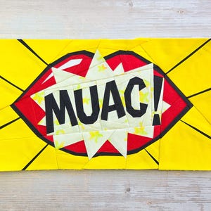 May include: A yellow fabric piece with a red lip design and the word "MUAC!" in black. The lips are outlined in black and set against a white and yellow starburst. The background has yellow rays.