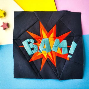 May include: A black fabric square with a red and orange starburst design. The starburst has the word "BAM!" written in blue letters.