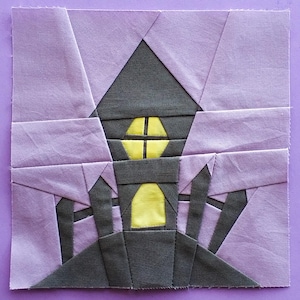 Puede incluir: Un bloque de colcha de retazos de tela morada y gris con un diseño de casa. La casa tiene una ventana amarilla y una puerta amarilla.