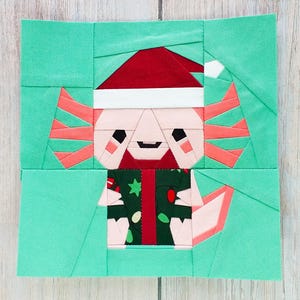 Peut inclure: Un bloc de tissu coloré représentant un axolotl festif. L'axolotl porte un bonnet de Père Noël et une veste verte avec un cadeau. Le fond est turquoise, et l'axolotl a des branchies roses et un corps rose clair. Thème de Noël.