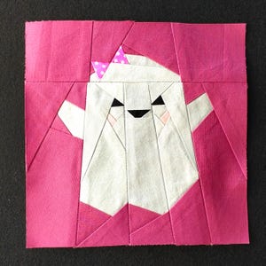 Puede incluir: Un fantasma blanco con un lazo rosa y ojos negros, sobre un fondo rosa. El fantasma está hecho de tela y es un diseño de patchwork.