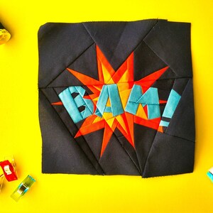 May include: A black fabric square with a red and orange starburst design and the word "BAM!" in blue letters. The fabric is surrounded by sewing supplies, including a spool of thread, a seam ripper, and fabric clips.