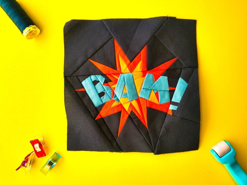 May include: A black fabric square with a red and orange starburst design and the word "BAM!" in blue letters. The fabric is surrounded by sewing supplies, including a spool of thread, a seam ripper, and fabric clips.