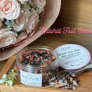 May include: A clear glass jar filled with a natural foot soak blend of dried rose petals, black tea, and sea salt. The jar has a white label with the text "LEtea NATURAL FOOT SOAK ROSE BLACK TEA" and a website address. The jar is sitting on a wooden tray with a spoon containing the foot soak.