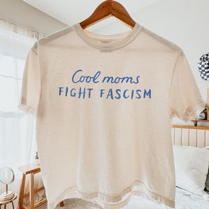 May include: A cream-colored t-shirt with the blue text "Cool moms FIGHT FASCISM." The short-sleeved shirt is hanging on a wooden hanger. The shirt is made of a soft, lightweight material.