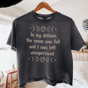 May include: A charcoal gray short-sleeve t-shirt featuring a light-colored graphic of moon phases and the text, In my defense, the moon was full and I was left unsupervised. The shirt is displayed on a wooden hanger against a bright indoor setting.