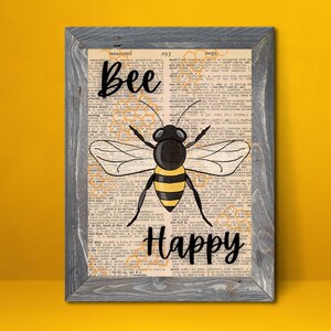 May include: Framed art print featuring a black and yellow bee illustration with the words "Bee Happy" in black script. The artwork is set against a vintage dictionary page background, framed in a gray wooden frame.