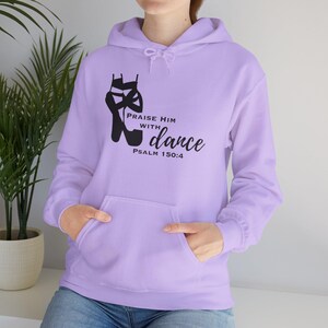 Puede incluir: Una sudadera con capucha de color lila claro con un bolsillo delantero y una capucha con cordón. La sudadera presenta un gráfico negro de una zapatilla de ballet y el texto "Praise Him with dance" con "Salmo 150:4" debajo.