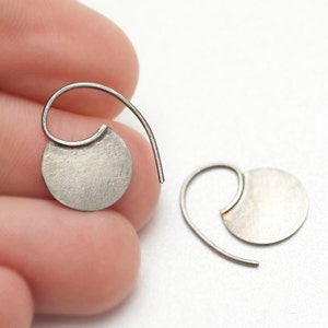 May include: A pair of silver-colored earrings. Each earring features a small, brushed metal disc attached to a curved wire. The earrings are designed to be worn through the ear, with a simple, minimalist style.