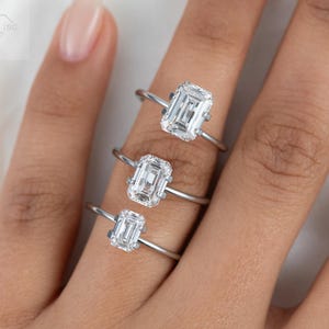 May include: Three silver rings with emerald cut diamonds of different sizes. The rings are stacked on a finger.