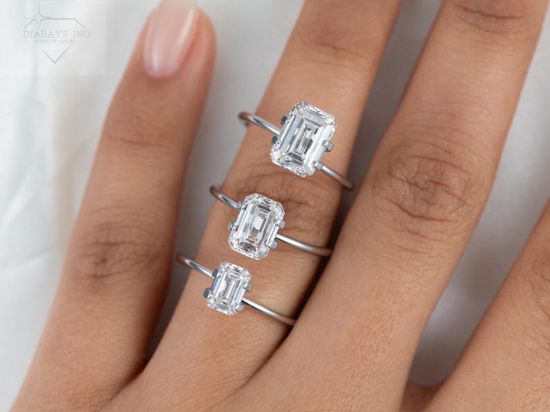 May include: Three silver rings with emerald cut diamonds of different sizes. The rings are stacked on a finger.